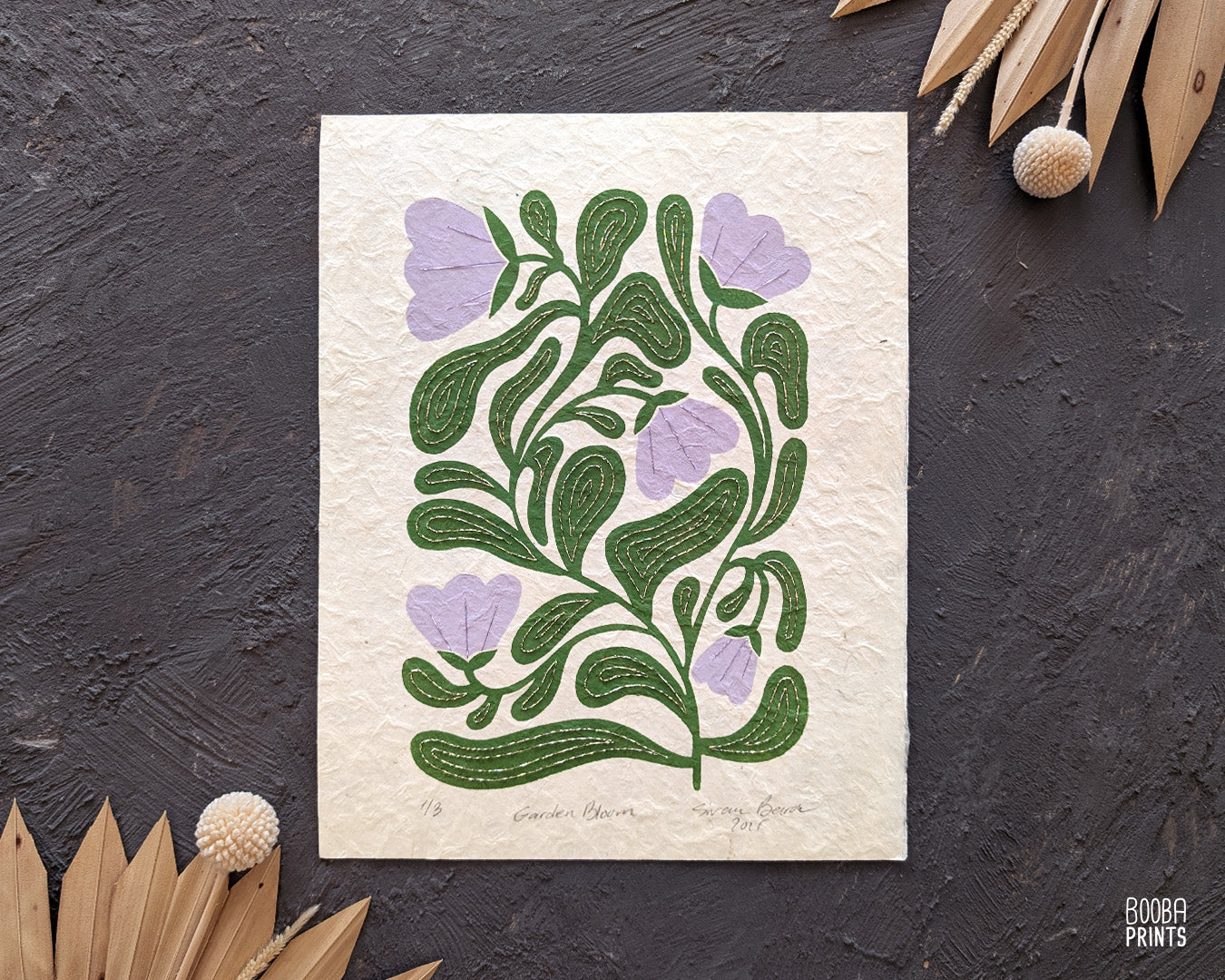 Handmade original Floral Print. Botanical design print on a textured Natural Lokta paper, placed against grey surface with dried flowers. Original linocut hand embroidered print in green and lavender, by Booba Prints