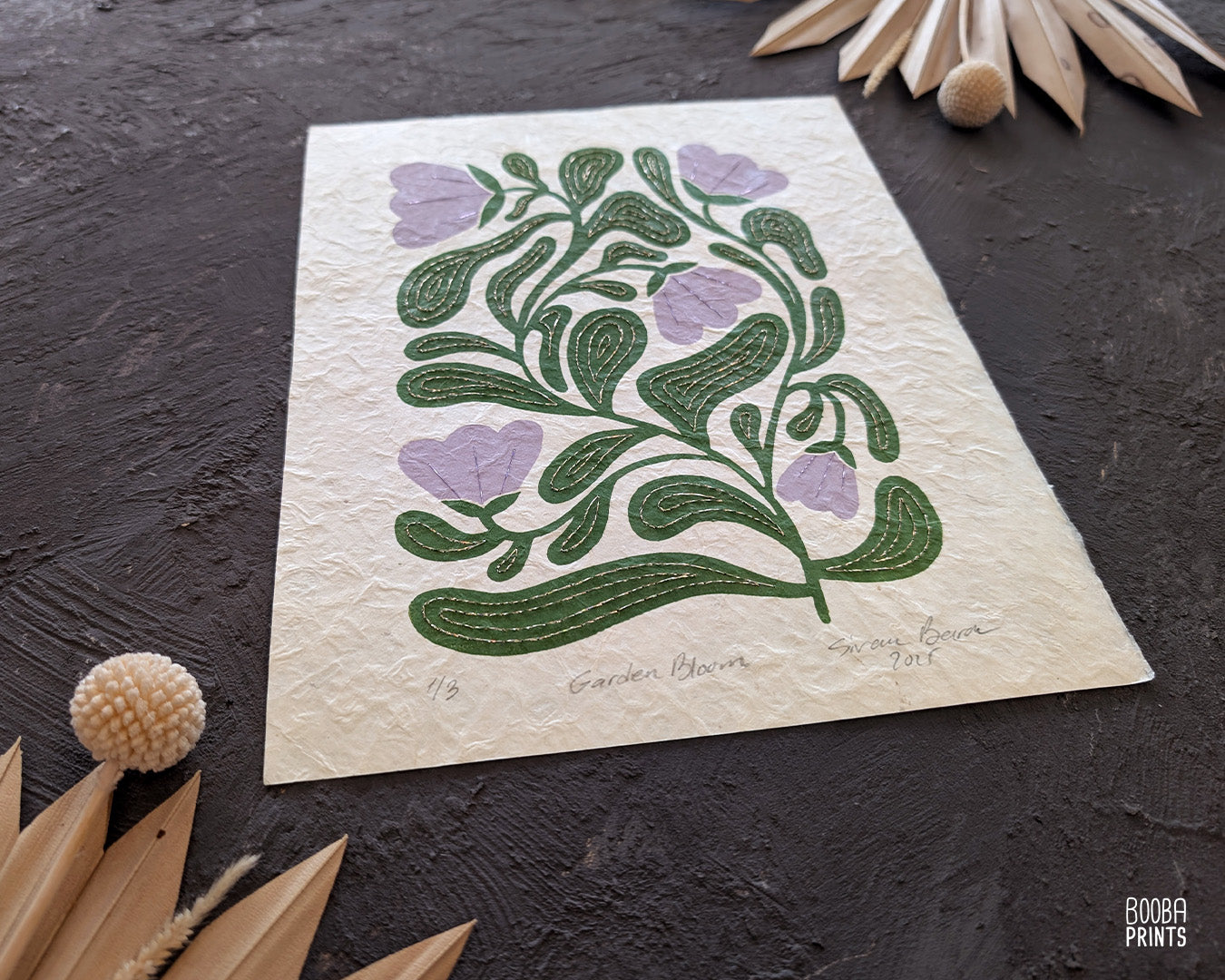 Handmade original Floral Print. Botanical design print on a textured Natural Lokta paper, placed against grey surface with dried flowers. Original linocut hand embroidered print in green and lavender, by Booba Prints