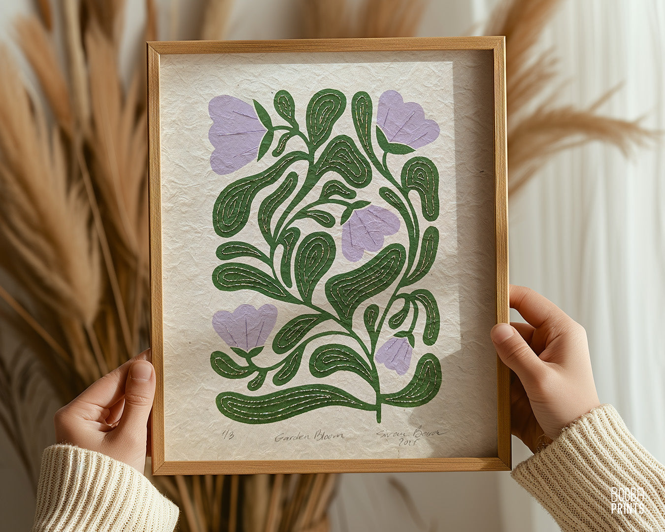 Hands holding a wooden picture frame of a green and lavender original linocut print. Hand printed and hand embroidered art piece. Unique mixed media wall art by Booba Prints