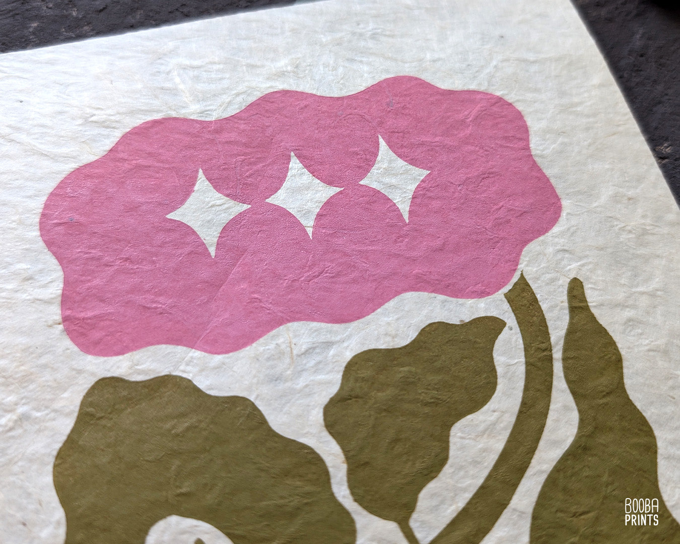 Close up of handmade linocut flower print, showing carved details and green and pink ink textures on natural Lokta paper. Art by Booba Prints