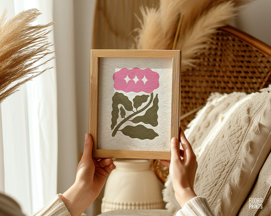Hands holding a framed mini linocut flower print in soft green and pink, minimal handmade wall art with botanical design. Art by Booba Prints