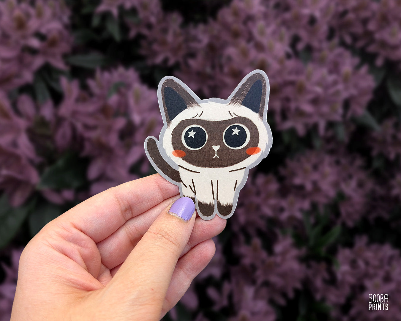 Hand holding a cute stylized Siamese cat sticker against a floral background. Adorable sticker for cat lovers. Waterproof vinyl sticker for planner, bottle, laptop.