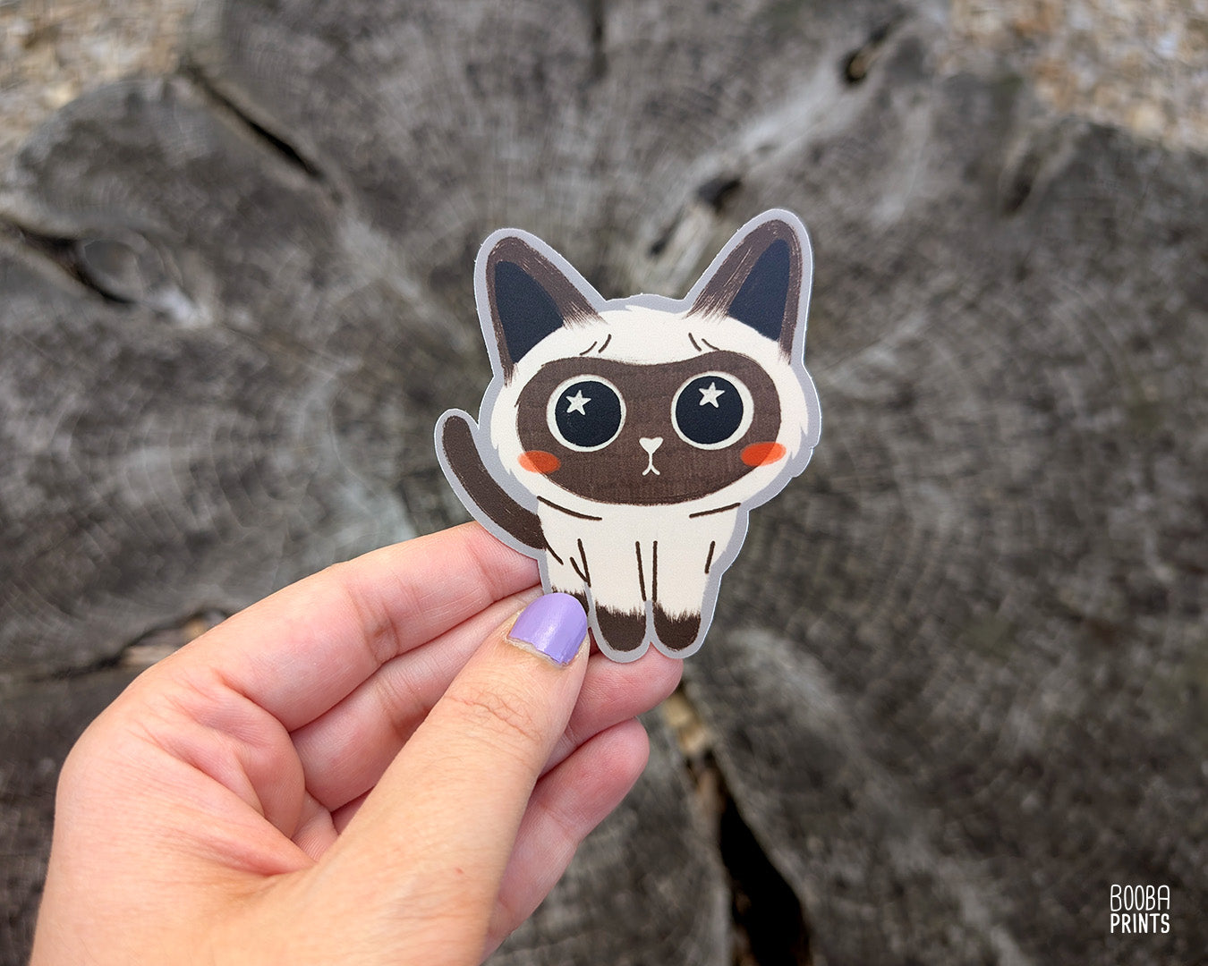 Hand holding a cute stylized Siamese cat sticker against a grey background. Adorable sticker for cat lovers. Waterproof vinyl sticker for planner, bottle, laptop.