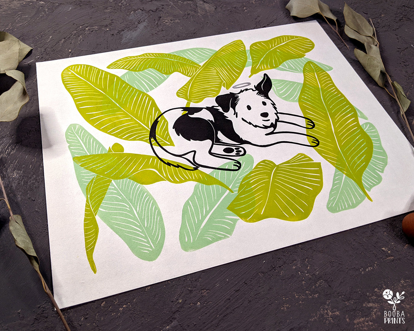 Original handmade linocut 4 color print of dog surrounded by banana leaves. Black and white dog with silver halo surrounded by 2 tones of green banana leaves .Dog lover gift. Dog print for home decor. Art by Booba Prints. Super Seconds Festiva. Online festival sale of seconds and samples, discounted prints, early holiday sale. UK festival.