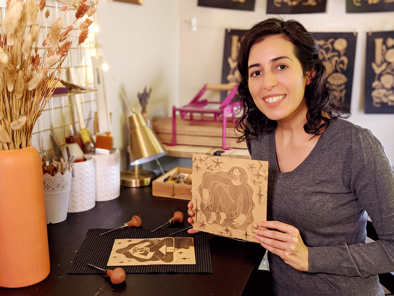 A picture of Booba Prints creator sitting at her work station, carving tools on the desk, holding a linoleum block featuring a woman design