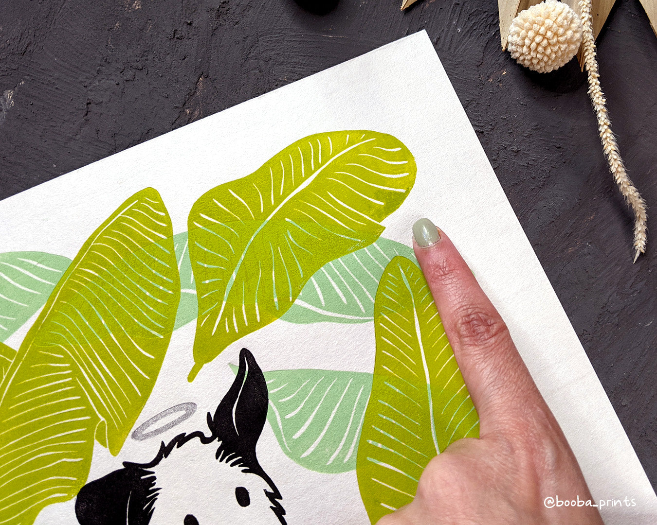 Original handmade linocut 4 color print of dog surrounded by banana leaves. Black and white dog with silver halo surrounded by 2 tones of green banana leaves .Dog lover gift. Dog print for home decor. Art by Booba Prints. Super Seconds Festiva. Online festival sale of seconds and samples, discounted prints, early holiday sale. UK festival.