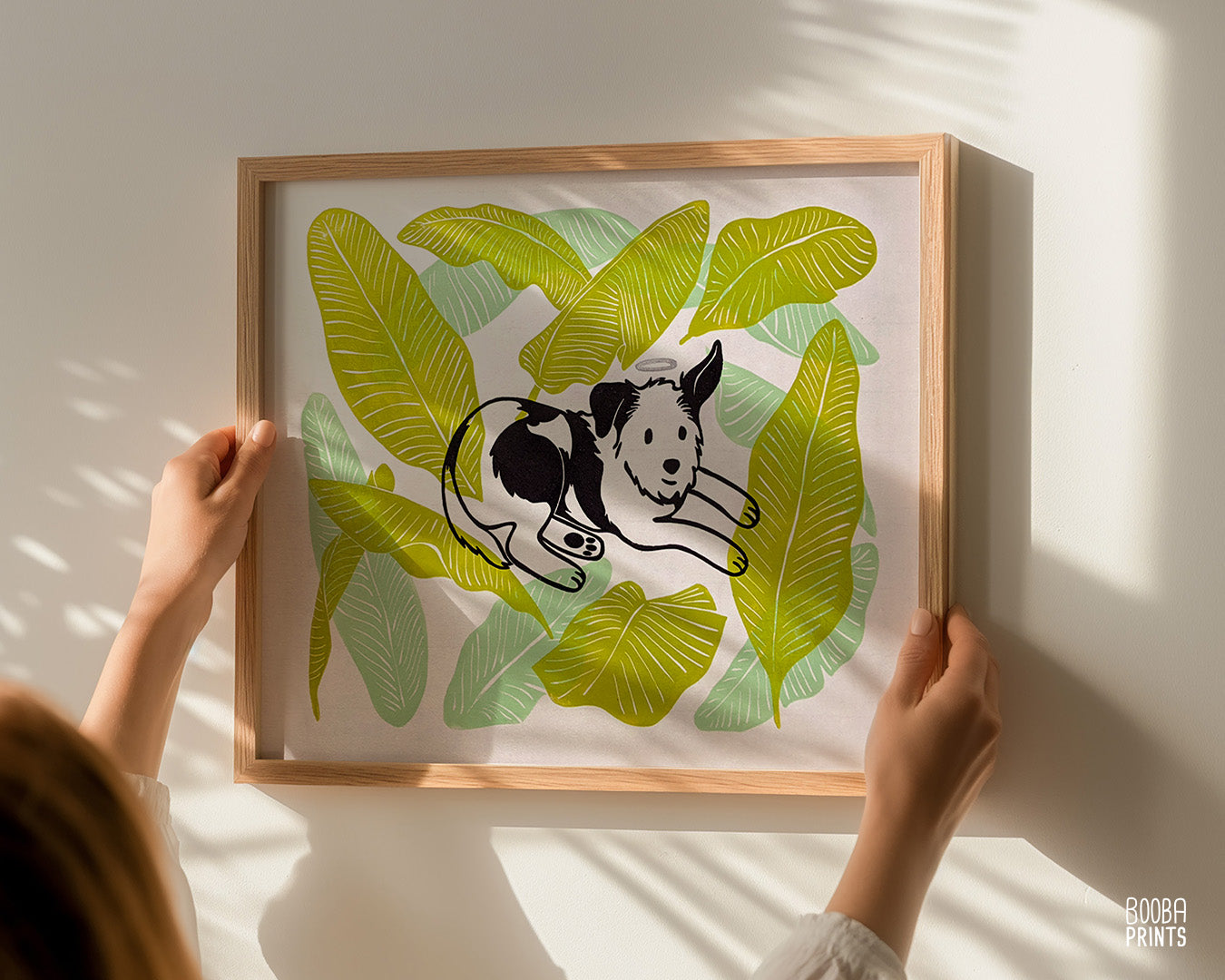 Original handmade linocut 4 color print of dog surrounded by banana leaves. Black and white dog with silver halo surrounded by 2 tones of green banana leaves .Dog lover gift. Dog print for home decor. Art by Booba Prints. Super Seconds Festiva. Online festival sale of seconds and samples, discounted prints, early holiday sale. UK festival.