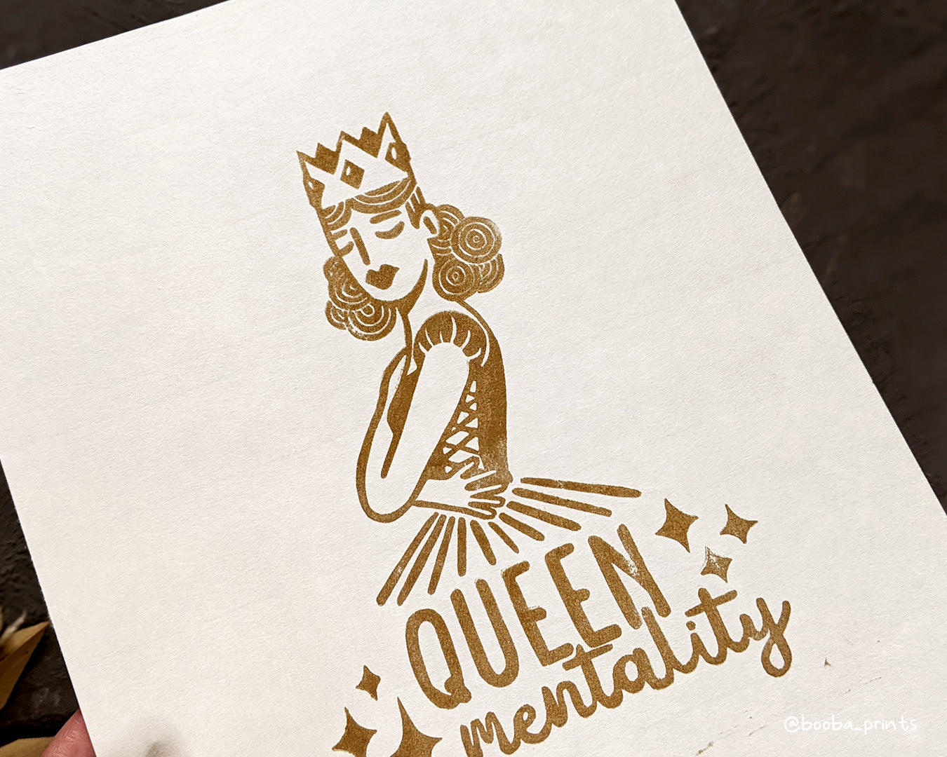 Handmade linocut print of Queen Mentality. Stylized illustration of a queen hugging herself and the text queen mentality. Printed with black or gold oil based ink on natural or Hosho paper. Empowering print for home decor. Super Seconds Festival, sale of samples and seconds. Artwork by Booba Prints.