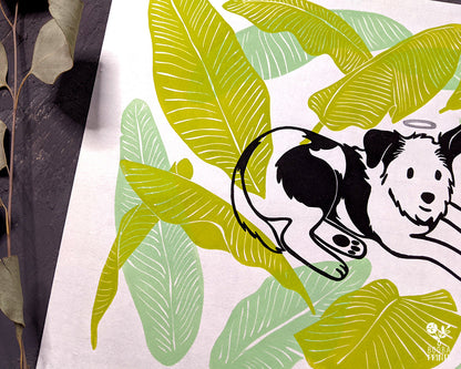 Original handmade linocut 4 color print of dog surrounded by banana leaves. Black and white dog with silver halo surrounded by 2 tones of green banana leaves .Dog lover gift. Dog print for home decor. Art by Booba Prints. Super Seconds Festiva. Online festival sale of seconds and samples, discounted prints, early holiday sale. UK festival.