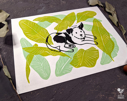 Original handmade linocut 4 color print of dog surrounded by banana leaves. Black and white dog with silver halo surrounded by 2 tones of green banana leaves .Dog lover gift. Dog print for home decor. Art by Booba Prints. Super Seconds Festiva. Online festival sale of seconds and samples, discounted prints, early holiday sale. UK festival.